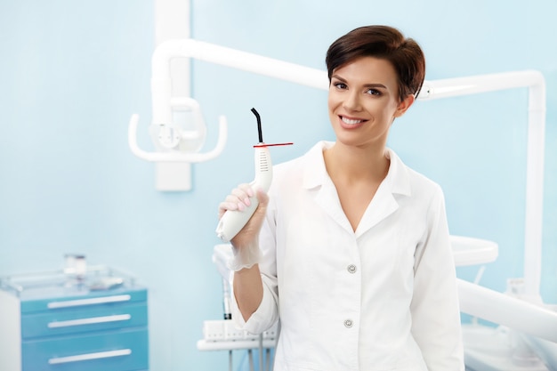 Jovem médico feminino no consultório do dentista. Mulher de sorriso bonita no jaleco branco que guarda a luz de cura dental azul. Clinica odontológica. Estomatologia