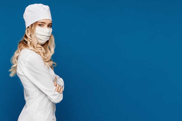 Jovem médico feminino no jaleco branco e máscara médica posando por um lado