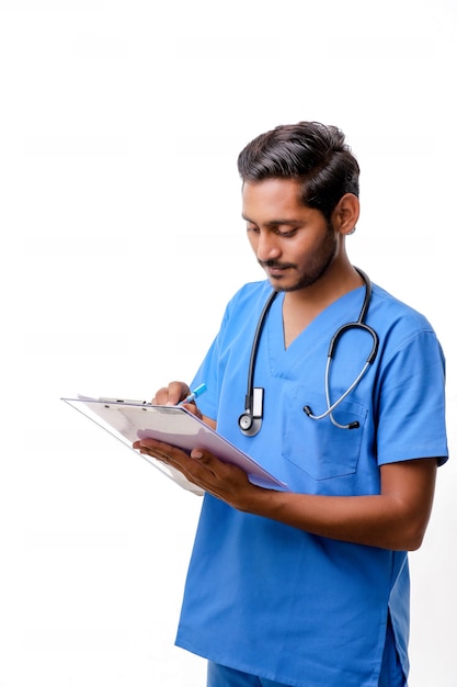 Jovem médico indiano masculino vestido de uniforme com estetoscópio fazendo anotações no bloco de notas isolado sobre fundo branco.