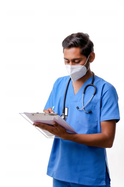 Jovem médico indiano masculino vestido de uniforme com estetoscópio fazendo anotações no bloco de notas isolado sobre fundo branco.