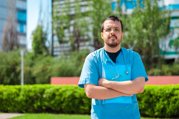 Jovem médico latino, de óculos, fora do hospital, braços cruzados, segurando uma pasta azul, com um estetoscópio no pescoço e olhando para a frente.