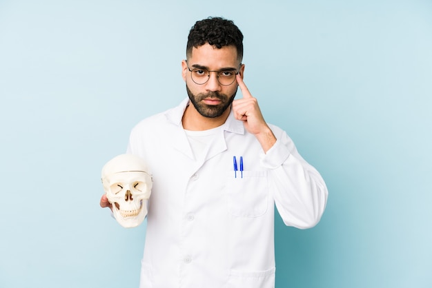 Jovem médico latino segurando um crânio isolado apontando o templo com o dedo, pensando, focado em uma tarefa.