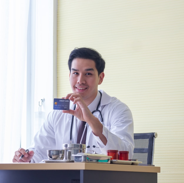 Jovem médico masculino asiático sorri mostrando cartão de crédito na mesa em um hospital