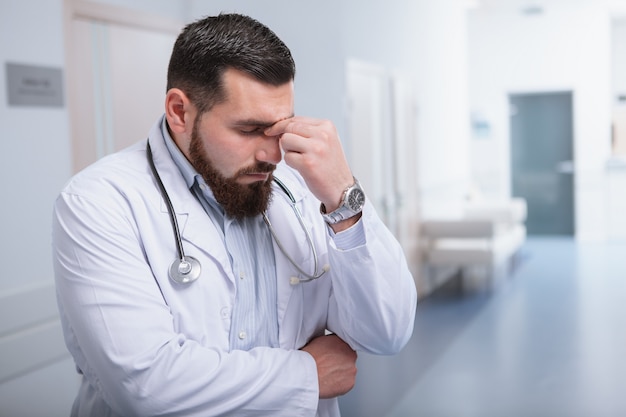 Jovem médico masculino, olhando cansado, em pé no corredor do hospital