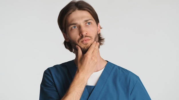 Foto jovem médico moreno mantendo a mão no queixo parecendo pensativo contra um fundo branco estagiário pensativo pensando sobre a operação de medicina futura