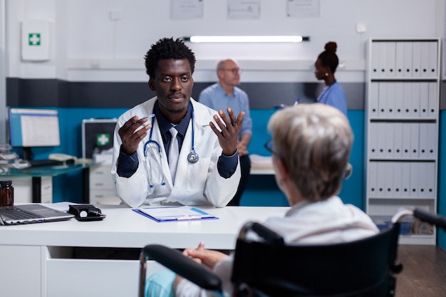 Jovem médico negro falando com paciente inválido na mesa do consultório