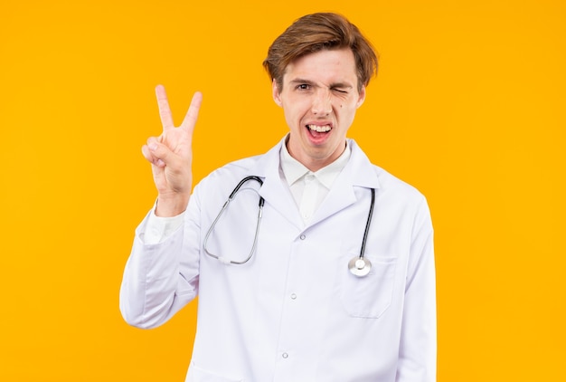 Jovem médico piscando alegre, vestindo túnica médica com estetoscópio, mostrando um gesto de paz isolado na parede laranja