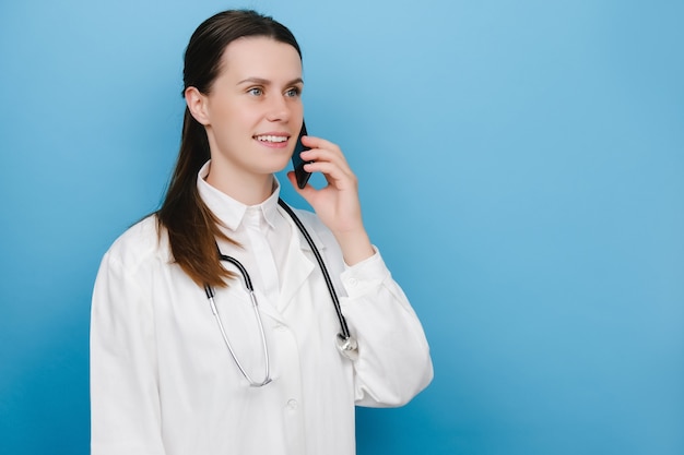 Jovem médico profissional feminino em uniforme médico branco e estetoscópio falando no celular, posando sobre a parede de fundo azul do estúdio. doc conversa. smartphone, conceito de tecnologia