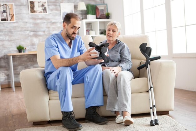 Jovem médico sentado no sofá no lar de idosos, ajudando a mulher idosa a usar óculos de realidade virtual.