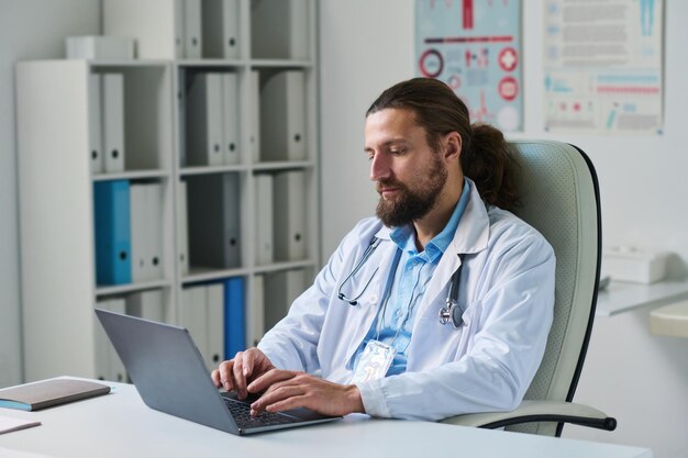Foto jovem médico sério digitando no teclado do laptop enquanto consulta o paciente on-line