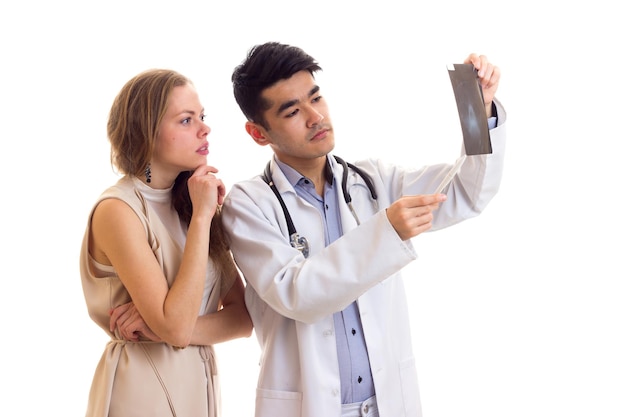 Jovem médico sério em vestido branco com estetoscópio mostrando raio-x para jovem e linda mulher