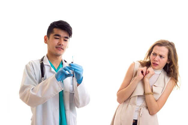 Jovem médico sorridente em vestido branco com estetoscópio se preparando para injeção de jovem assustada