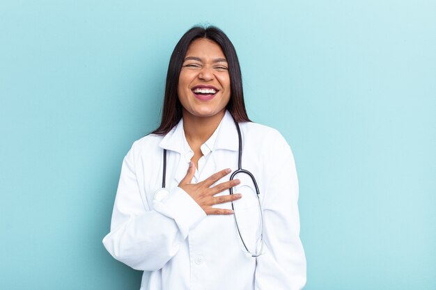 Jovem médico venezuelano isolado sobre fundo azul ri alto, mantendo a mão no peito.