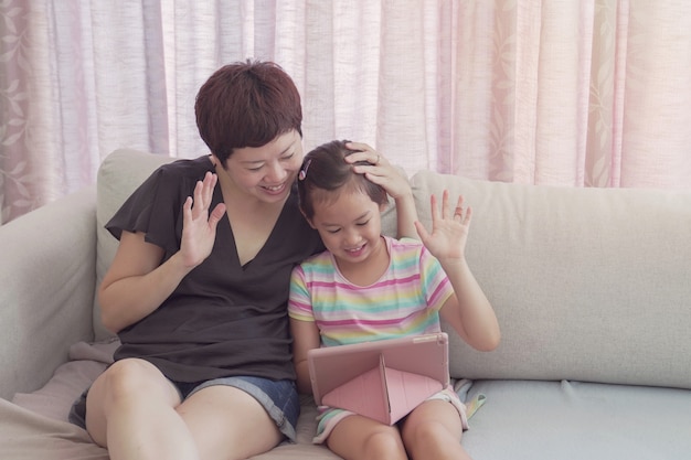 Jovem menina asiática e sua mãe fazendo chamadas facetime por vídeo com o laptop em casa, usando o aplicativo on-line para aprendizagem com zoom, distanciamento social, isolamento, educação em casa, aprendendo remotamente o conceito