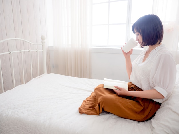 Jovem menina asiática lendo um livro e bebendo na cama.