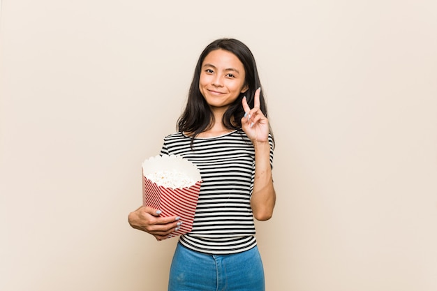Jovem menina asiática segurando um balde de pipoca, mostrando o número dois com os dedos.