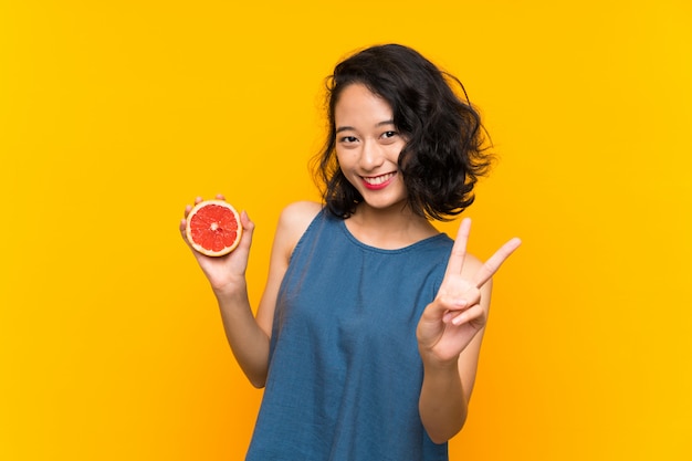 Jovem menina asiática segurando uma toranja sobre parede laranja isolada, sorrindo e mostrando sinal de vitória