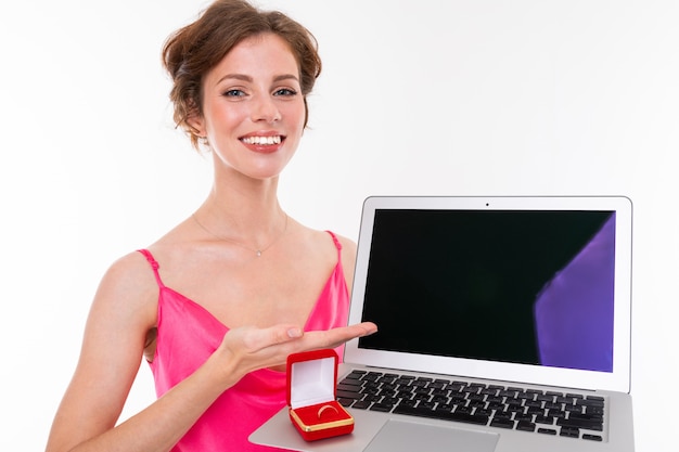 Jovem menina bonita com cabelos remendados castanhos ondulados, pele limpa, dentes planos, sorriso lindo, em jersey rosa, detém uma caixa de anel de treinamento e laptop