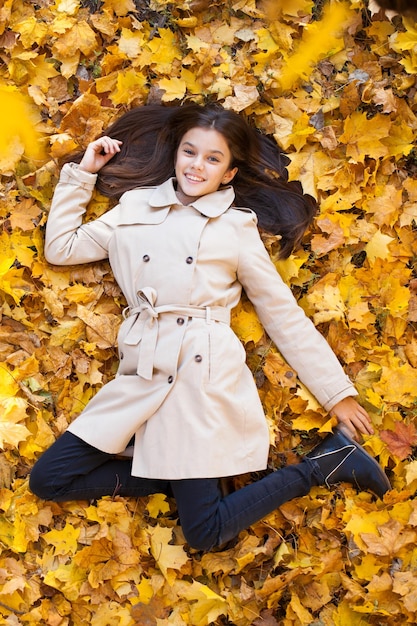 Foto jovem menina bonita em casaco bege deitada em folhas amarelas vista de cima no parque de outono