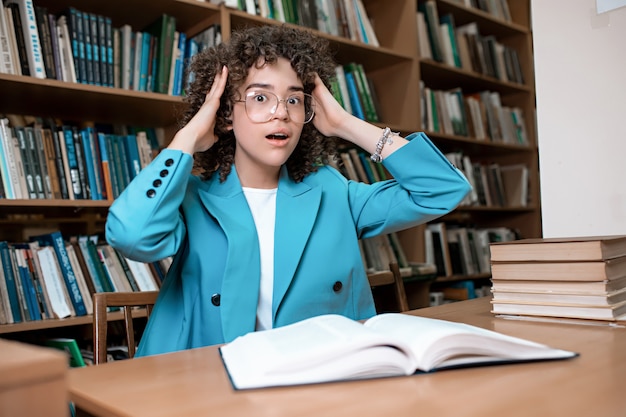 Jovem menina bonita encaracolada em copos sentado na biblioteca