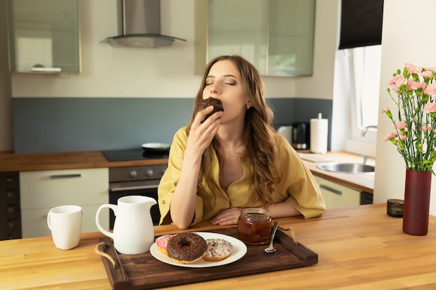 Jovem menina bonita está tomando café da manhã em casa na cozinha. Ela bebe o café da manhã e come um bolinho de chocolate.