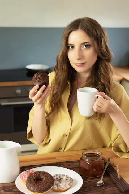 Foto jovem menina bonita está tomando café da manhã em casa na cozinha. ela bebe o café da manhã e come um bolinho de chocolate.