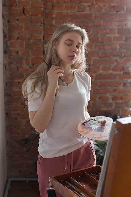 Jovem menina bonita loira com pincel, misturando cores na paleta