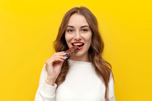 Foto jovem menina come e morde grande barra de chocolate e sorri e se alegra em fundo isolado amarelo