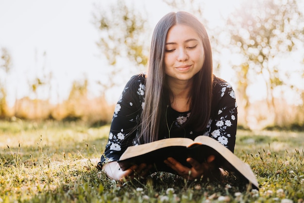 Jovem menina crente deitada na grama e estudando a bíblia, no campo ao pôr do sol. Copiar espaço