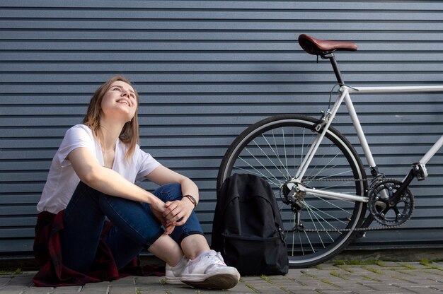 Jovem menina elegante senta-se perto da bicicleta e mochila e sonhos, sorri contra a parede cinza