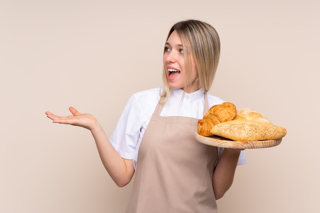 Jovem menina loira com avental. Padeiro feminino segurando uma mesa com vários pães com expressão facial de surpresa