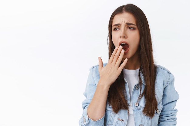 Jovem menina morena cansada e entediada com jaqueta jeans, olhando carrancuda para longe e bocejando, cobrir a boca aberta, preciso dormir, acordar cedo quero uma xícara de café, fundo branco de pé