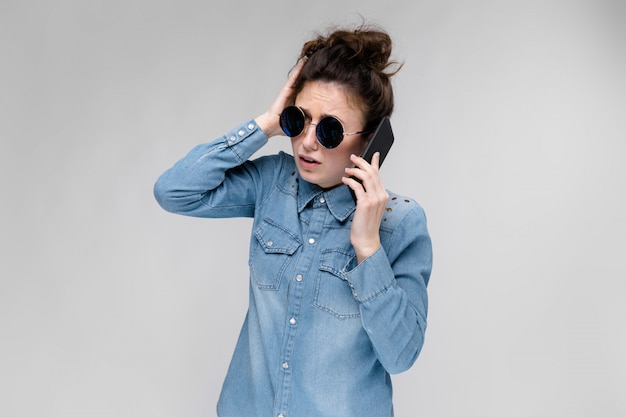 Jovem menina morena de óculos redondos. Os cabelos são reunidos em um coque. Menina com um telefone preto. A garota está falando ao telefone.