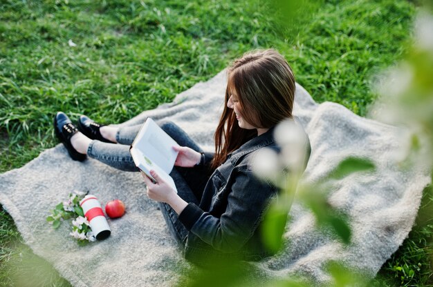 Jovem menina morena em jeans, sentado na manta contra a árvore de flor de primavera e ler o livro.