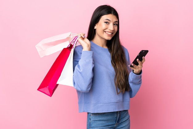 Jovem menina morena isolada rosa segurando sacolas de compras e escrever uma mensagem com o celular para um amigo