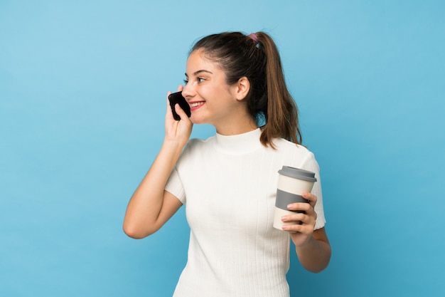 Jovem menina morena isolado azul segurando café para levar e um celular