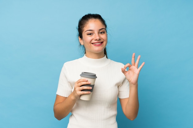 Jovem menina morena isolado azul segurando café para tirar enquanto faz sinal de OK