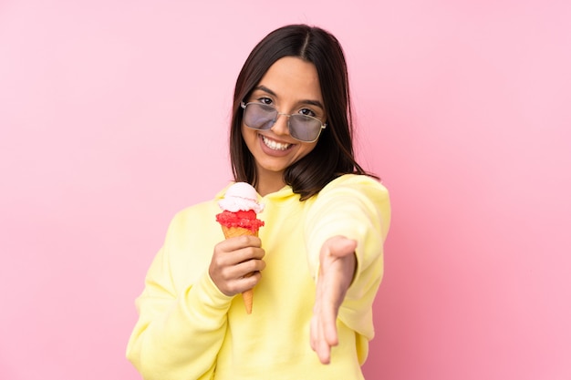 Jovem menina morena segurando um sorvete de corneta sobre parede rosa isolada, apertando as mãos para fechar um bom negócio