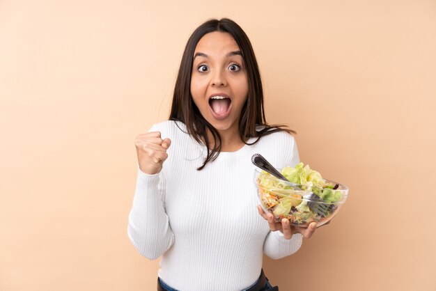 Jovem menina morena segurando uma salada sobre parede isolada, comemorando uma vitória na posição de vencedor