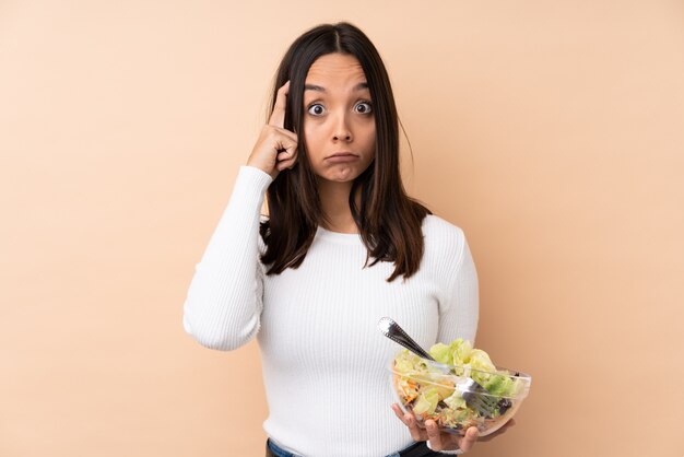 Jovem menina morena segurando uma salada sobre parede isolada, pensando em uma idéia