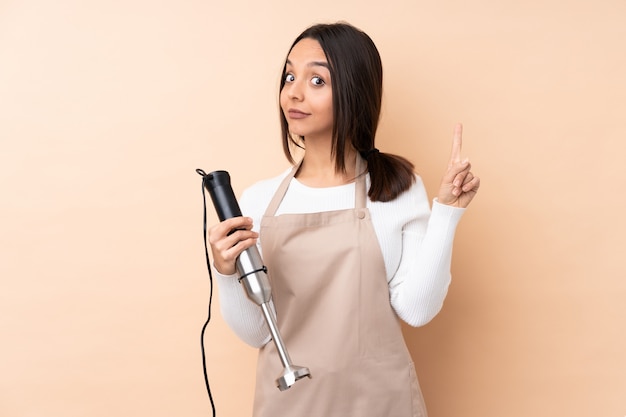 Jovem menina morena usando liquidificador mão sobre parede isolada, mostrando e levantando um dedo em sinal dos melhores