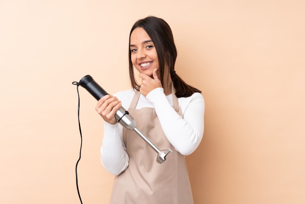 Jovem menina morena usando liquidificador mão sobre parede isolada, olhando para o lado