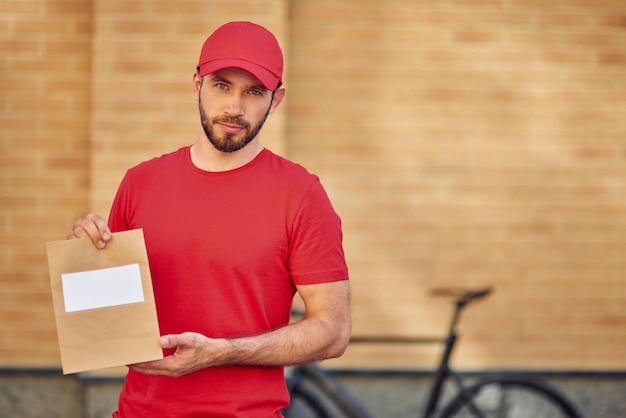 Jovem mensageiro masculino caucasiano de uniforme vermelho segurando um pequeno saco de papel com espaço de cópia para