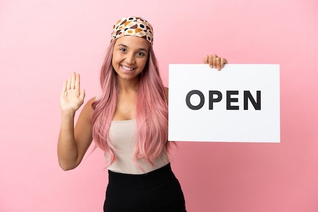 Jovem mestiça com cabelo rosa isolada em um fundo rosa segurando um cartaz com o texto ABRIR e saudando