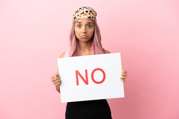 Jovem mestiça com cabelo rosa isolada em um fundo rosa segurando um cartaz com o texto NÃO com expressão triste