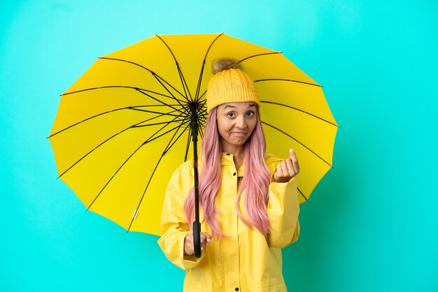 Jovem mestiça com casaco à prova de chuva e guarda-chuva fazendo gestos de dinheiro