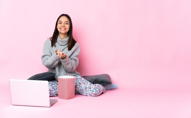 Jovem mestiça comendo pipoca enquanto assiste a um filme no laptop segurando copyspace imaginário na palma da mão para inserir um anúncio