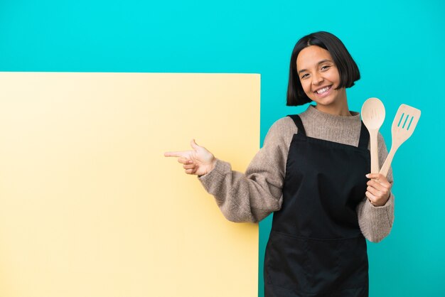 Jovem mestiça cozinheira com um grande cartaz isolado na parede azul apontando o dedo para o lado e apresentando um produto