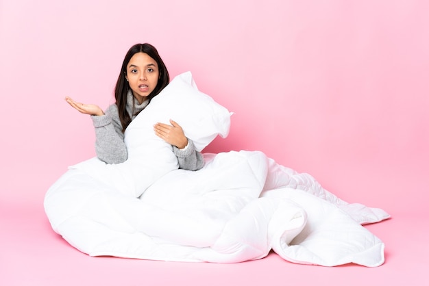 Jovem mestiça de pijama sentada no chão fazendo gesto de dúvida