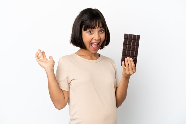 Jovem mestiça grávida isolada em um fundo branco tomando uma pastilha de chocolate e surpreendendo